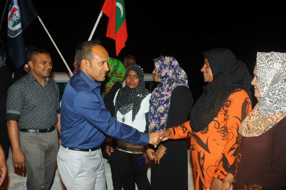 Dr Jameel in an open-necked shirt shaking hands with a line of women at an outdoor gathering, the national flag behind.